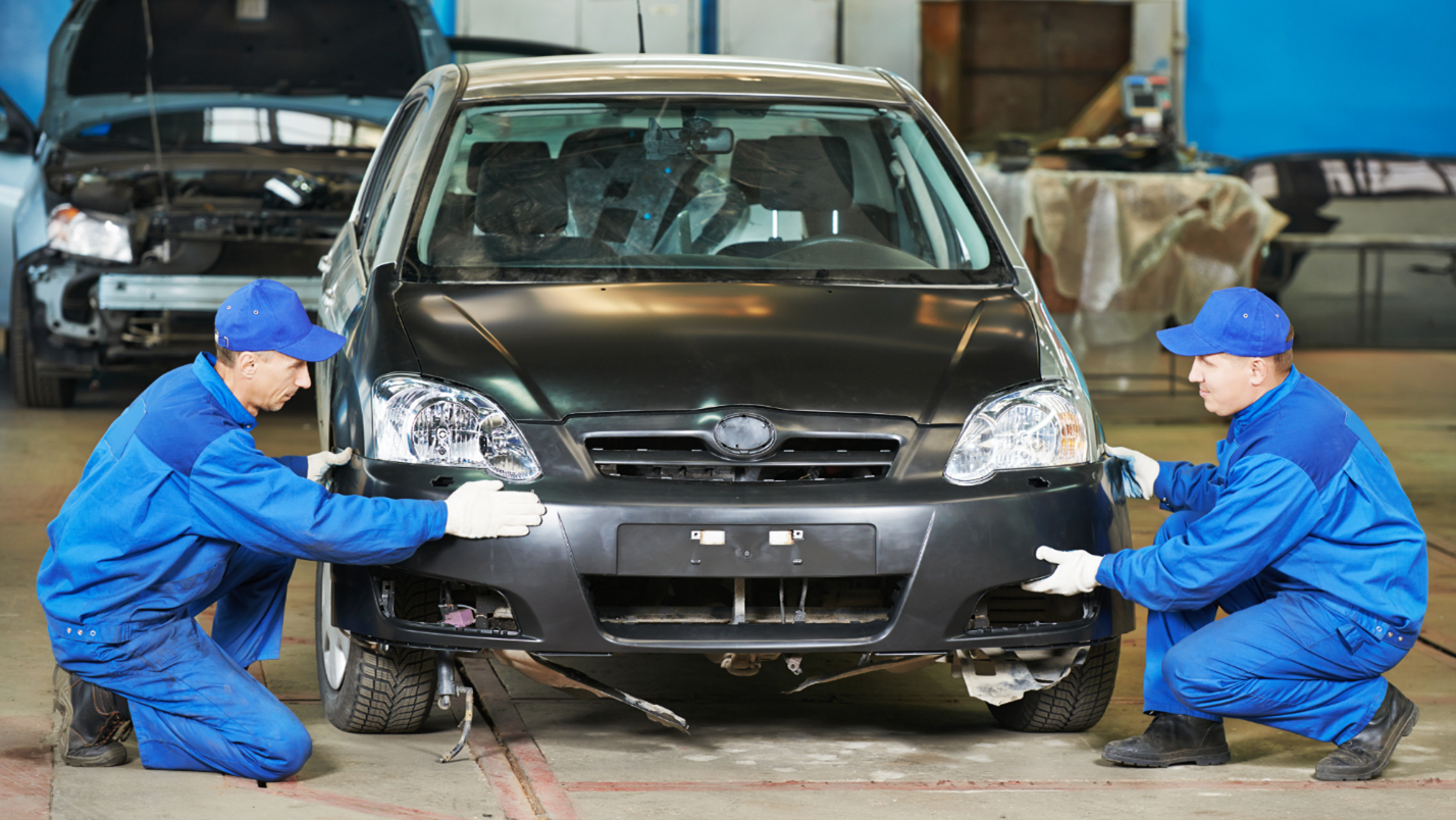 Two men in blue jumpsuits replace the bumper of a small black car.