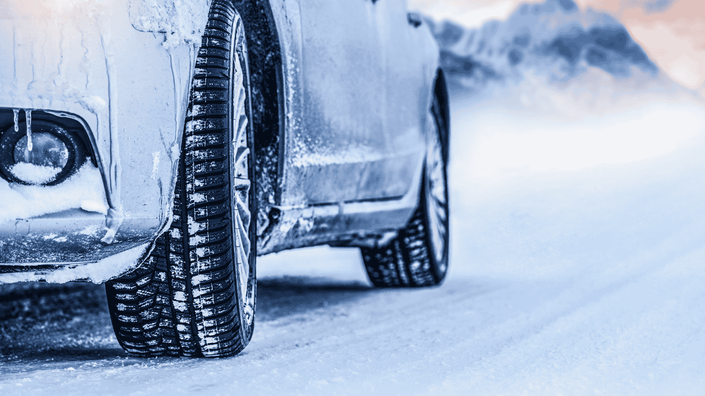 A close up of a car’s lower body and tires as it drives in the snow.