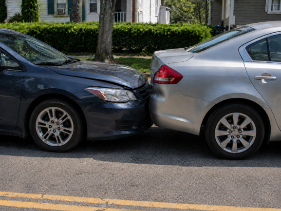 Dark sedan with dented hood rear-ends a silver sedan on a suburban street, showing minor fender-bender damage.