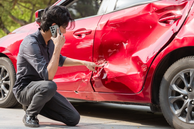 Man kneels beside a red car with severe side-door dents while holding a phone and pointing at collision damage.