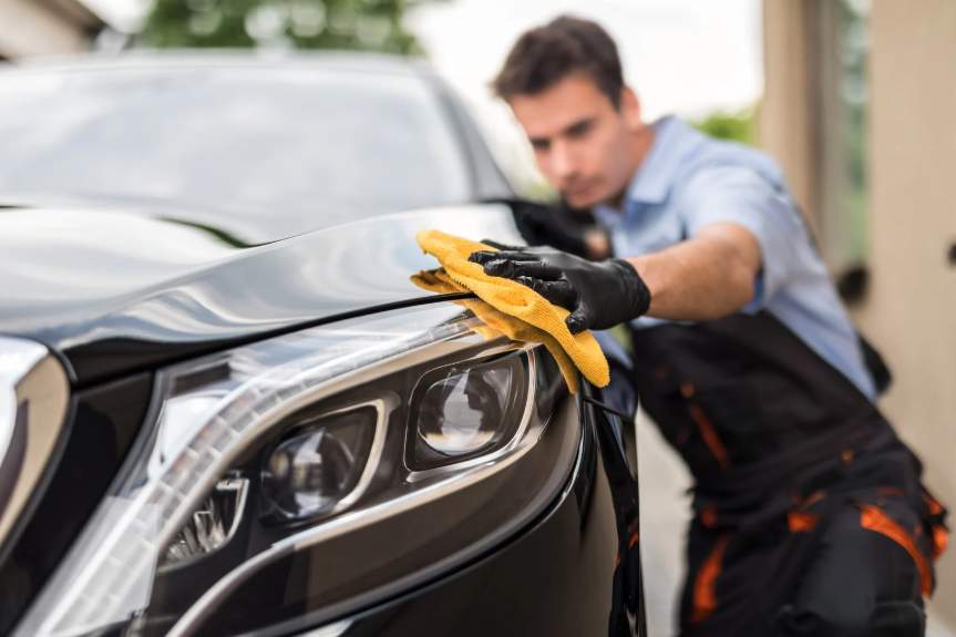 Auto detailer in black gloves wipes a glossy black car hood with a bright yellow microfiber cloth beside a sleek headlight.